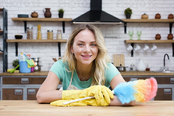 Smiling housekeeper leaning kitchen counter holding feather duster looking camera