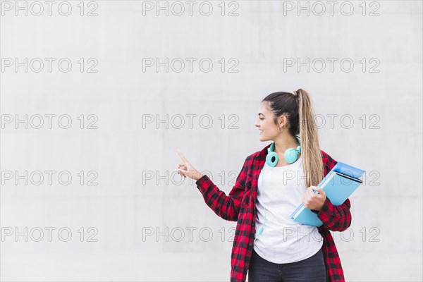 Portrait smiling female university student holding books hand pointing her finger white wall with copy space