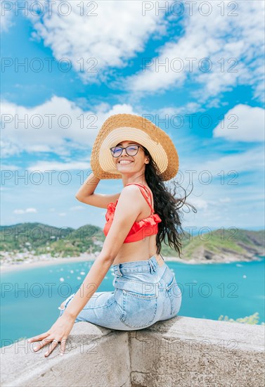 Portrait of tourist girl in a hat at a viewpoint with a beach in the background. Happy tourist female sitting at a viewpoint with beach in the background