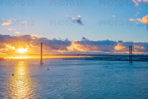 View of 25 de Abril Bridge famous tourist landmark of Lisbon connects Lisboa and Almada on Setubal Peninsula over Tagus river with tourist yacht silhouette at sunset and flying plane. Lisbon