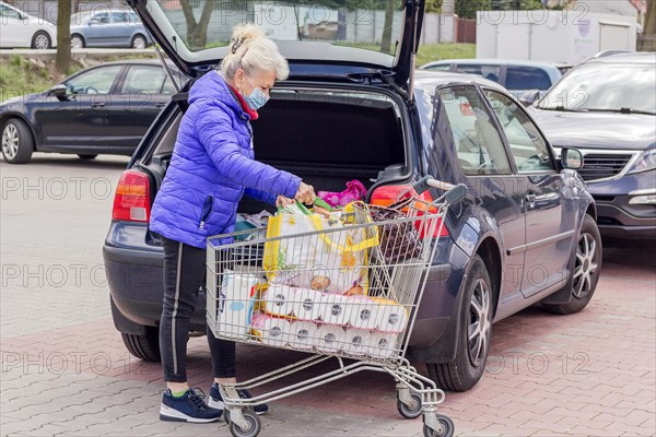 A mature woman wearing protective mask loads purchases into a car in the parking lot. Everyday life during coronavirus pandemic. Shopping concept