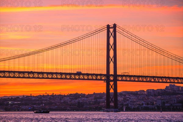 View of 25 de Abril Bridge famous tourist landmark of Lisbon connecting Lisboa and Almada over Tagus river with tourist boat silhouette at sunset. Lisbon