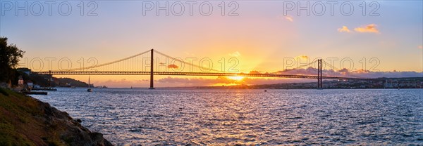 Panorama of 25 de Abril Bridge famous tourist landmark of Lisbon connecting Lisboa and Almada on Setubal Peninsula over Tagus river with tourist yacht silhouette at sunset. Lisbon