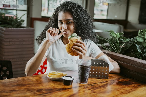 Latin woman sucking her fingers holding a hamburger in a restaurant. Portrait of an afro girl enjoying hamburger in a restaurant