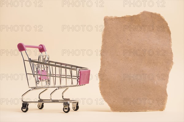 Empty miniature shopping trolley near torn paper piece against colored backdrop