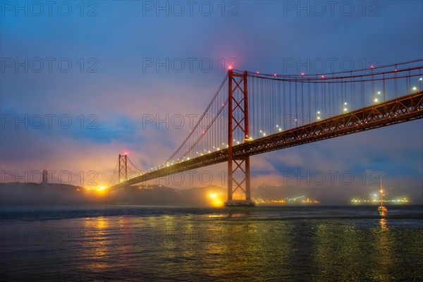 View of 25 de Abril Bridge famous tourist landmark of Lisbon connecting Lisboa and Almada in heavy fog mist wtih yacht boats passing under. Lisbon