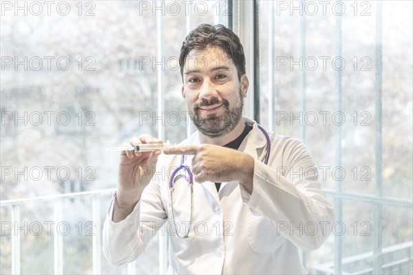 Handsome hispanic man wearing doctor uniform and stethoscope at medical clinic smiling to the camera while presenting a vaccine