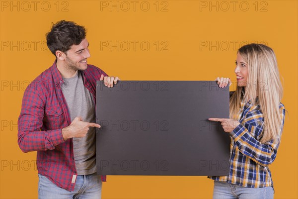 Portrait young couple pointing their finger blank black placard against orange backdrop