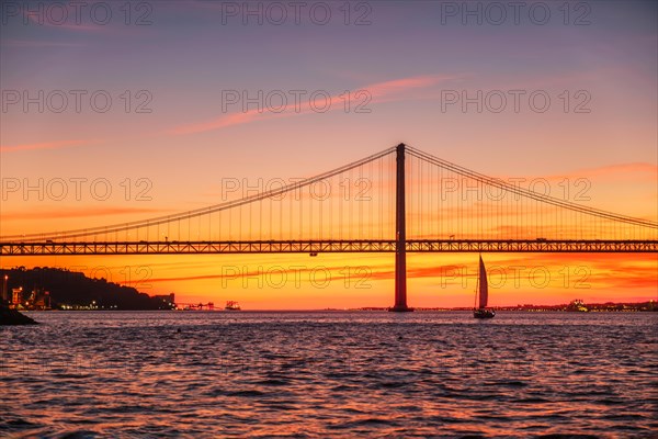 View of 25 de Abril Bridge famous tourist landmark of Lisbon connecting Lisboa and Almada over Tagus river with tourist yacht silhouette at sunset. Lisbon