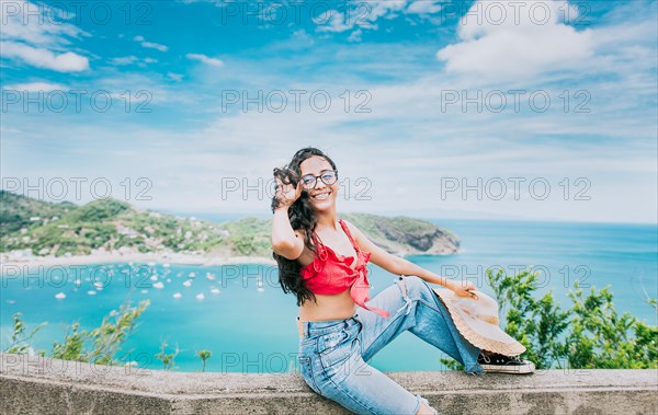 Happy female tourist sitting at a viewpoint with beach in the background. Lifestyle of a tourist girl with a hat in a viewpoint with a beach in the background