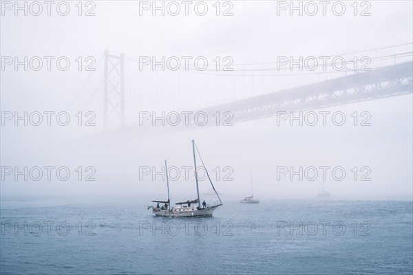 View of 25 de Abril Bridge famous tourist landmark of Lisbon connecting Lisboa and Almada in heavy fog mist wtih yacht boats passing under. Lisbon