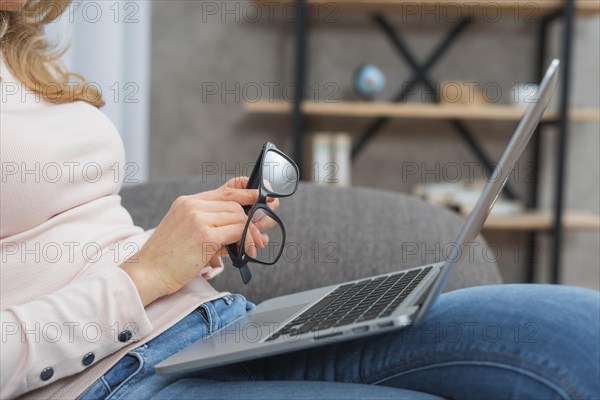 Woman holding eyeglasses hand sitting sofa with open laptop her lap