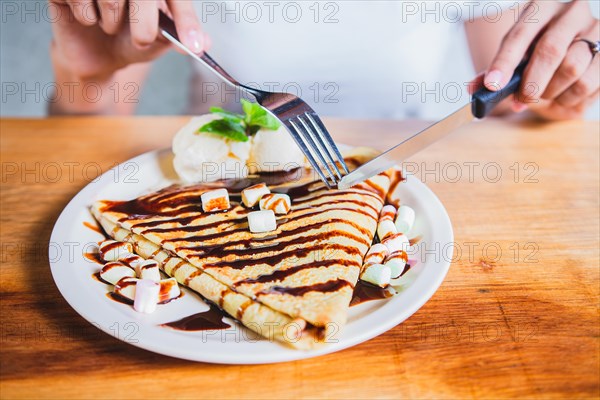 Hands of person with fork cutting chocolate crepe and ice cream. Woman eating chocolate crepe and ice cream with fork