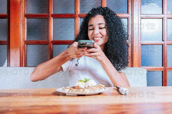 Young woman using cell phone and taking a picture of chocolate crepe. Happy girl photographing a plate of chocolate crepe and ice cream