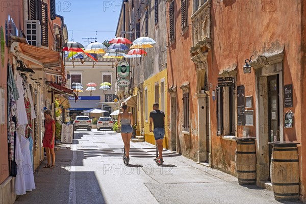 Tourists on e-scooters riding through shopping street with colourful umbrellas in the historic town centre of Novigrad