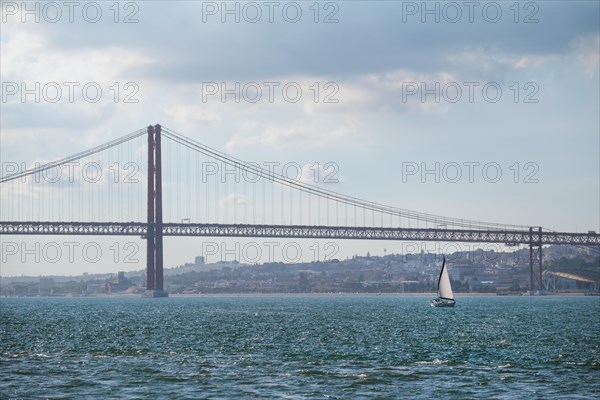 View of 25 de Abril Bridge famous tourist landmark of Lisbon connecting Lisboa and Almada over Tagus river with tourist yacht boat. Lisbon