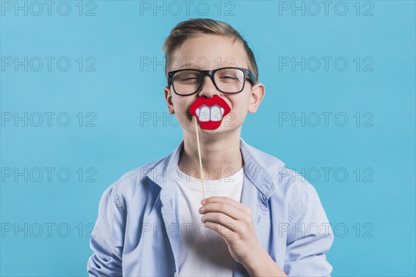 Boy wearing black eyeglasses holding smiling prop front his mouth against blue background