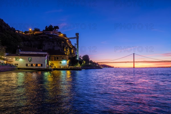 View of 25 de Abril Bridge famous tourist landmark of Lisbon over Tagus river and Boca do Vento Elevator and seaside restaurants with people in the evening. Lisbon