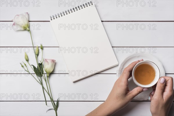 Person holding tea cup with spiral notepad beautiful flowers wooden desk