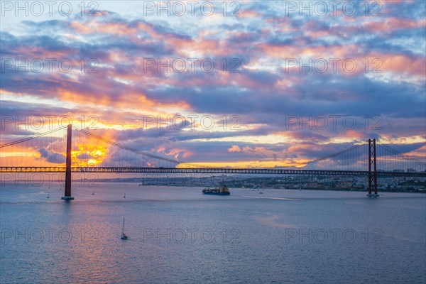 View of 25 de Abril Bridge famous tourist landmark of Lisbon over Tagus river with tourist yacht boats and cargo container ship on sunset. Lisbon