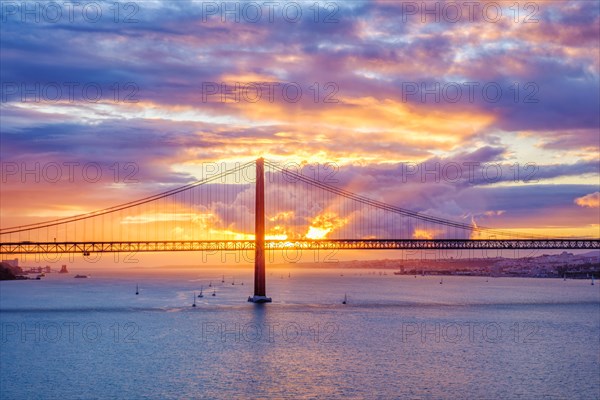 View of 25 de Abril Bridge famous tourist landmark of Lisbon connecting Lisboa and Almada on Setubal Peninsula over Tagus river with tourist yacht boats on sunset. Lisbon