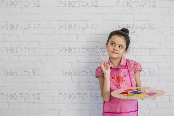 Portrait girl with paintbrush palette standing against white wall