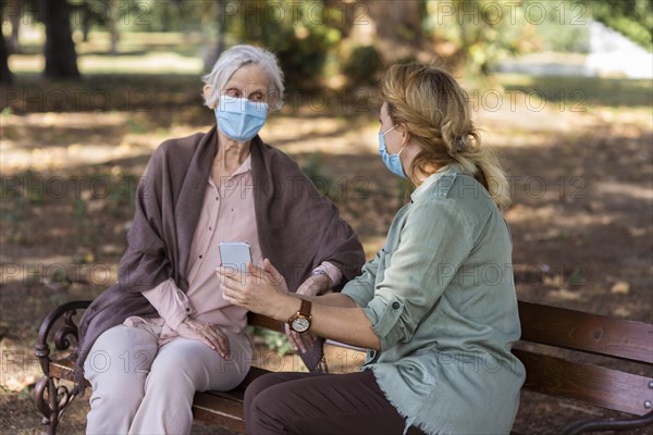 Elder woman conversing with woman bench outdoors while holding smartphone