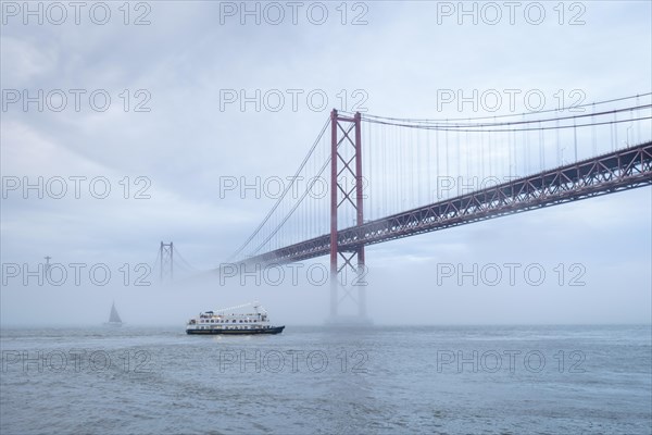 View of 25 de Abril Bridge famous tourist landmark of Lisbon connecting Lisboa and Almada in heavy fog mist wtih yacht boats passing under. Lisbon
