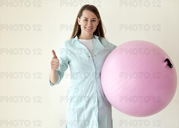 Smiley physiotherapist holding exercise ball giving thumbs up