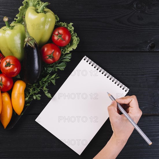 Human hand writing spiral notepad with fresh vegetables black wooden backdrop