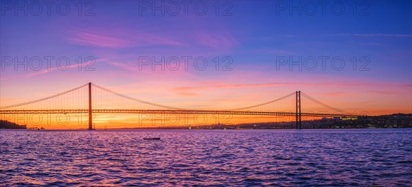 Panorama of 25 de Abril Bridge famous tourist landmark of Lisbon connecting Lisboa and Almada over Tagus river with tourist yacht silhouette at sunset. Lisbon