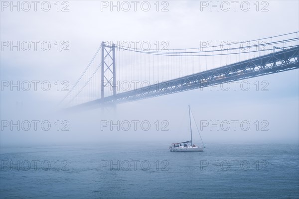 View of 25 de Abril Bridge famous tourist landmark of Lisbon connecting Lisboa and Almada in heavy fog mist wtih yacht boats passing under. Lisbon