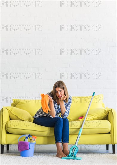 Sad young woman sitting yellow sofa looking orange rubber gloves