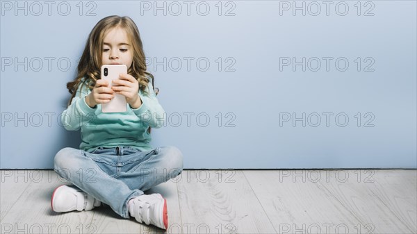 Portrait little girl sitting hardwood floor looking smartphone