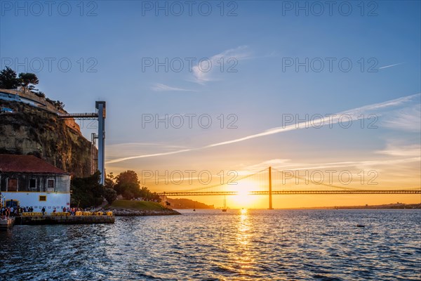 View of 25 de Abril Bridge famous tourist landmark of Lisbon over Tagus river and Boca do Vento Elevator and seaside restaurants with people at the sunset. Lisbon