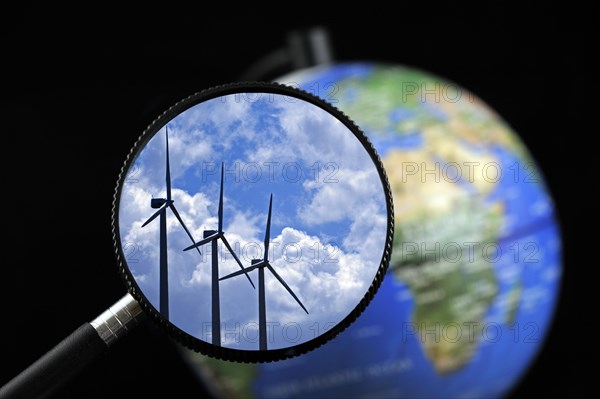 Wind farm turbines against cloudy sky seen through magnifying glass held against illuminated terrestrial globe
