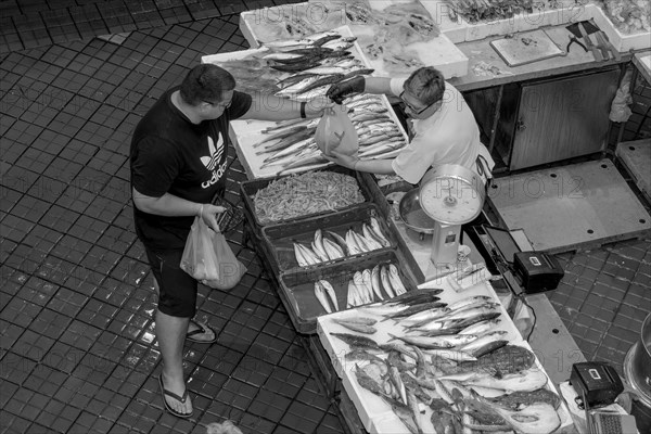 Black and white bird's eye view photo of a stall full of fish with the seller and a buyer