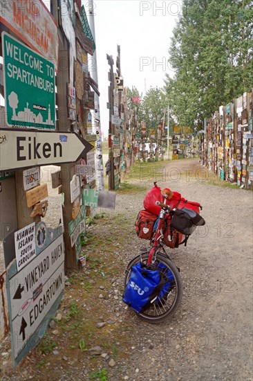 Various traffic and place signs face a bicycle loaded with luggage