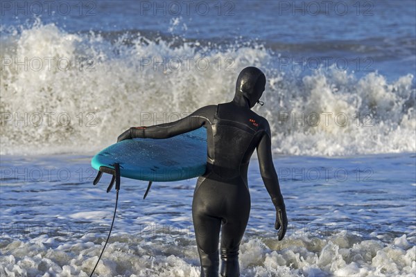 Surfer in black wetsuit on the beach entering sea water with surfboard to ride the waves