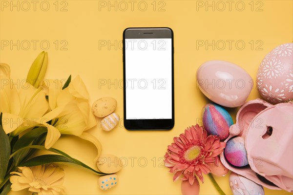 White blank screen mobile phone decorated with lily gerbera flower colorful easter eggs yellow background