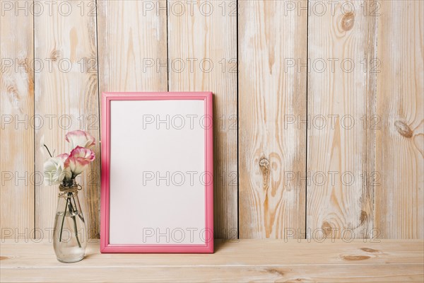 Flower vase near white blank frame with pink border against wooden wall