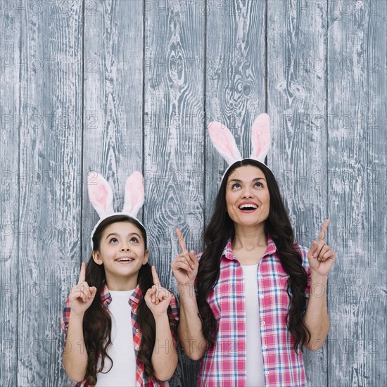 Excited mother daughter with bunny ears pointing finger upward against wooden backdrop