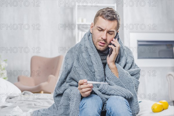 Young man sitting bed wrapped scarf looking thermometer talking mobile phone