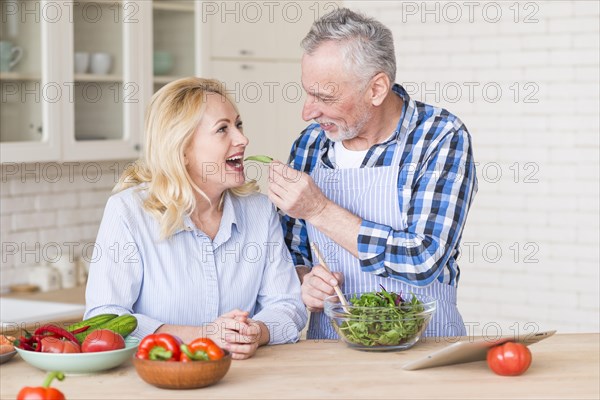 Smiling senior man feeding fresh green salad her wife kitchen