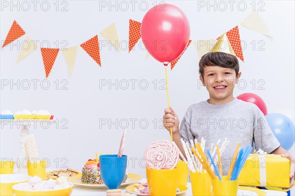 Smiling boy wearing party hat holding balloon gift standing variety food table