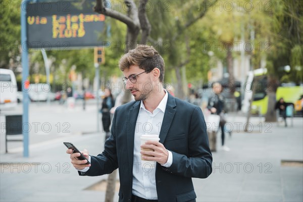 Young businessman holding disposable coffee cup looking mobile phone
