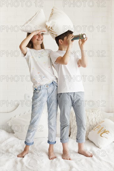 Eye shielding girl standing bed with her brother looking through telescope