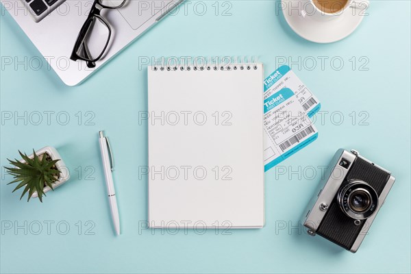 Blank spiral notepad with air tickets surrounded with laptop eyeglasses pen camera coffee cup blue desk