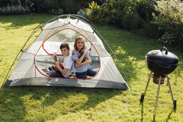 Boy playing ukulele sitting back back his sister tent near barbecue grill