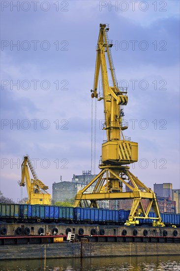 River port crane loading open-top gondola cars on cloudy day. Empty river drag boats or barges moored by pier. Empty cars ready for loading. Vertical image